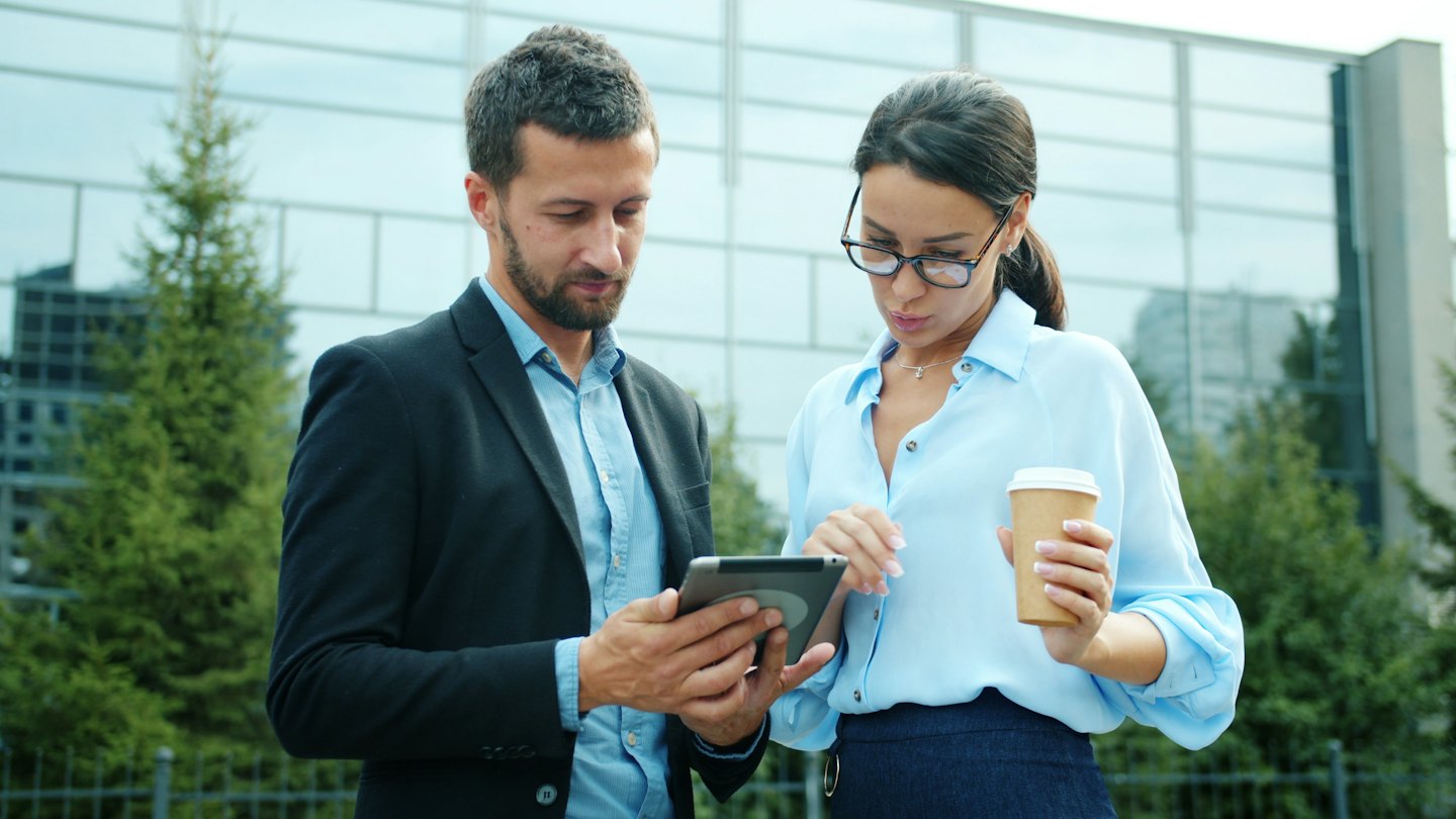 Business professionals discussing a tablet outdoors near a glass building.